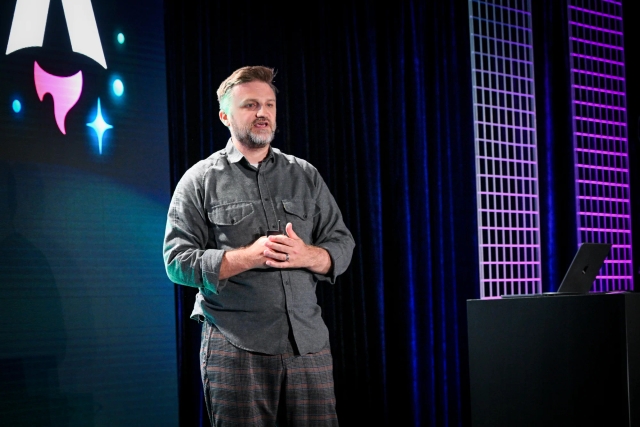 Matthew Phillips speaking on stage next to a podium with a laptop on it. Behind him is the Astro Together logo on-screen.