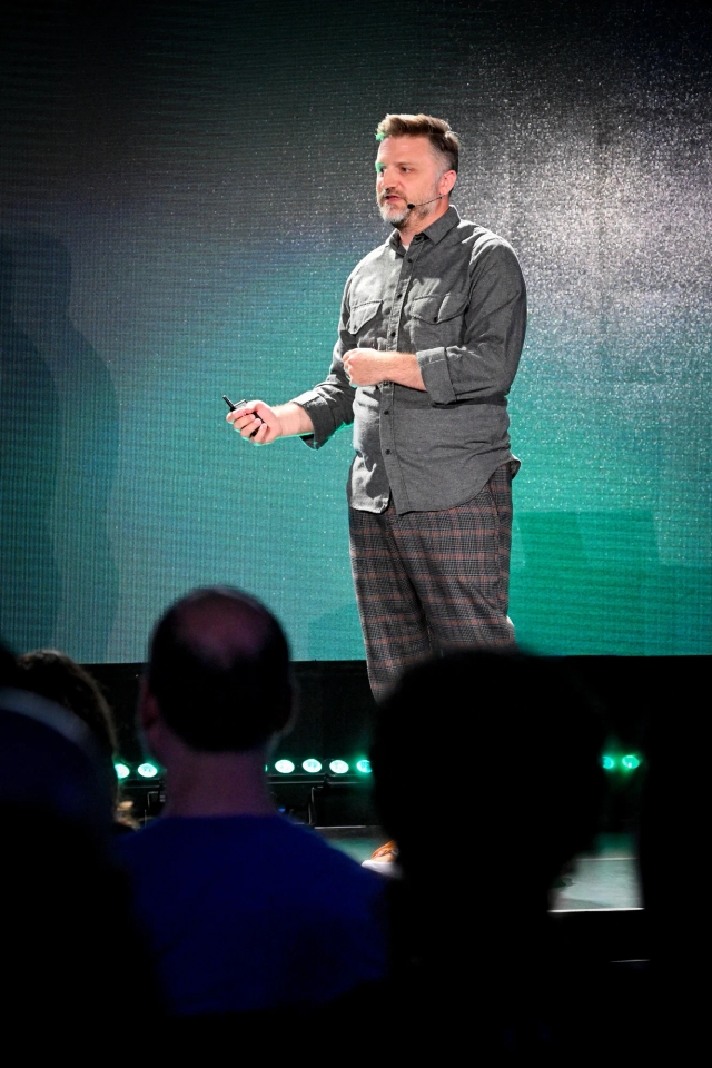 Portrait view of Matthew Phillips speaking on stage with audience members in the foreground.