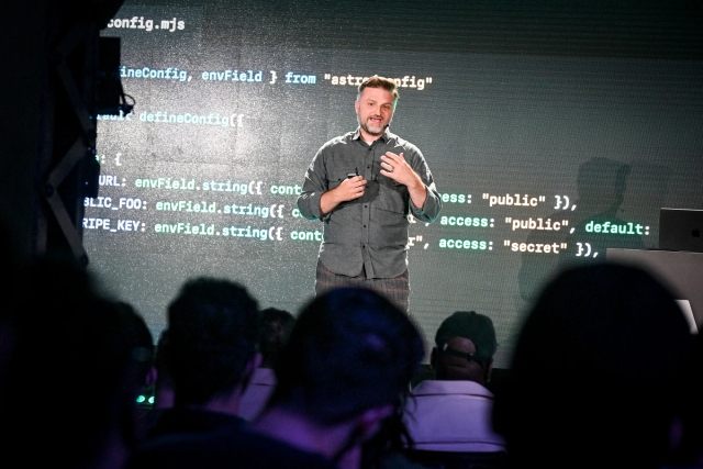 View from the audience of Matthew Phillips speaking on stage, standing in front of a code example.