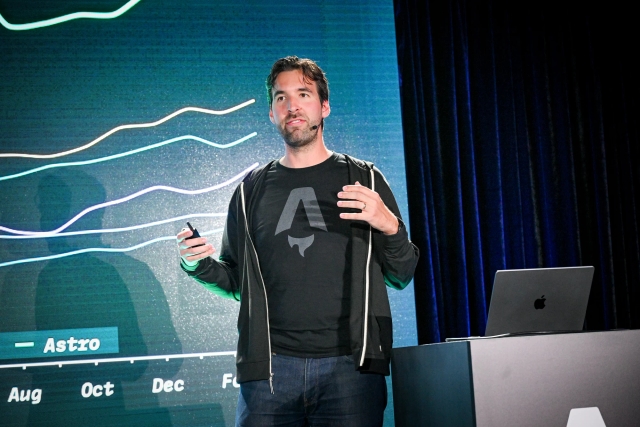 Fred K. Schott speaking on stage next to a podium with a laptop on it. Behind him is a slide depicting a graph.