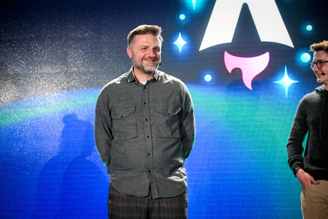 Closeup of Matthew Phillips standing with other keynote speakers on stage. His hands are behind his back and he is smiling at the audience. Off to the right, Ben Holmes is smiling at him.